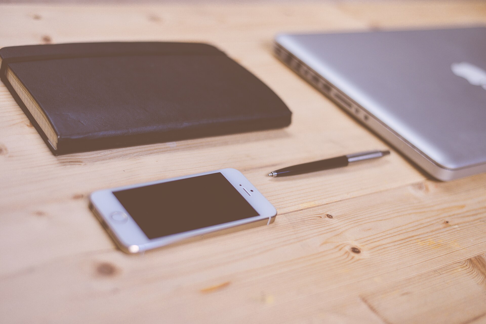 A phone, notebook, pen, and laptop on a light wooden desk.